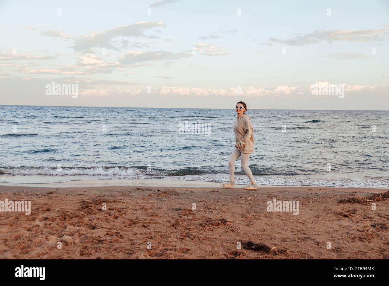 Giovane donna felice con felpa e leggings in avorio e occhiali da sole bianchi che corre sulla spiaggia guardando la macchina fotografica facendo un volto divertente. Splendido concetto di natura Foto Stock