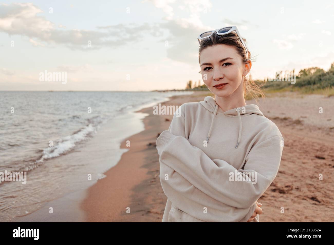 Giovane donna sottile con abiti in avorio e occhiali da sole bianchi in piedi sulla spiaggia incrociate le mani vicino al lago guardando la fotocamera godendo di una splendida vista. Esterno Foto Stock