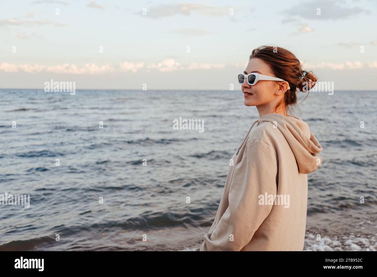 Giovane donna snella con abiti in avorio e occhiali da sole bianchi in piedi sulla spiaggia incrociati con le mani sul lago per ammirare gli schizzi d'acqua e godersi una splendida vista. Ou Foto Stock