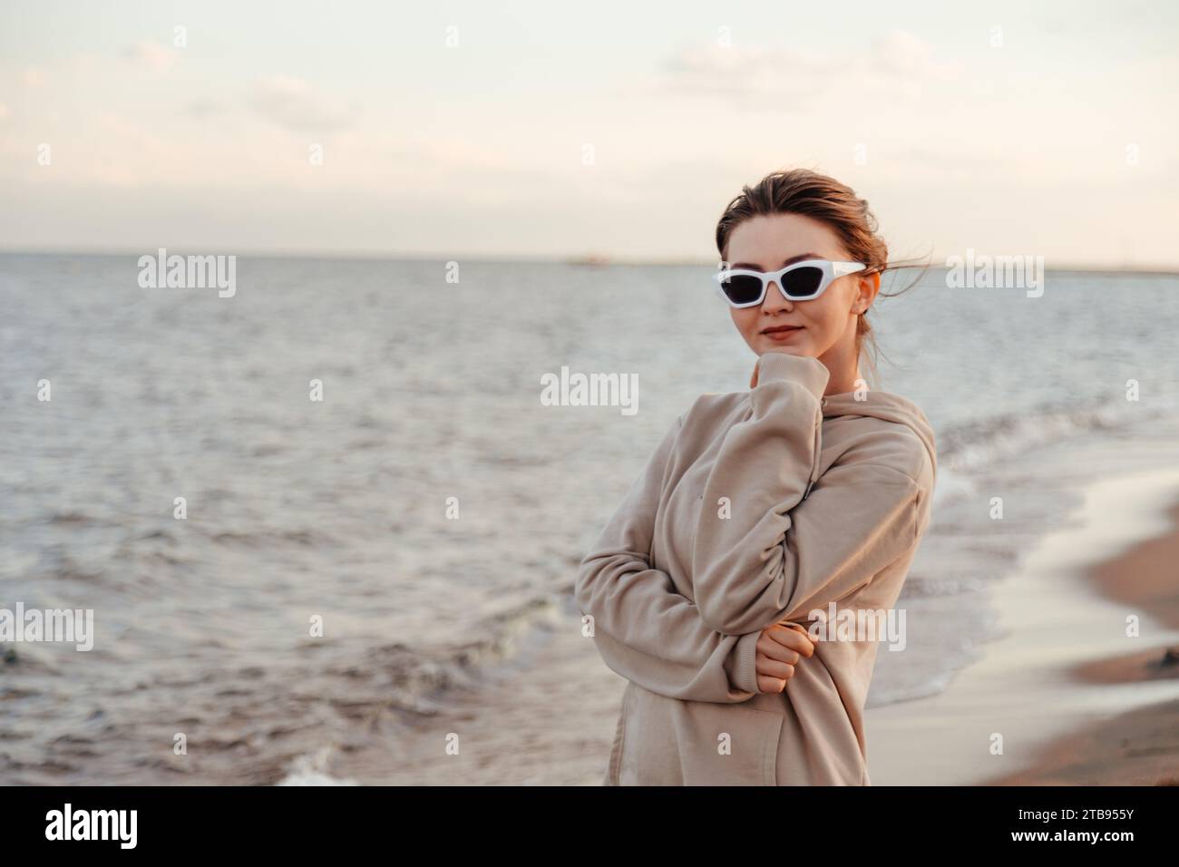 Giovane donna sottile con abiti in avorio e occhiali da sole bianchi in piedi sulla spiaggia incrociate le mani vicino al lago guardando la fotocamera godendo di una splendida vista. Esterno Foto Stock