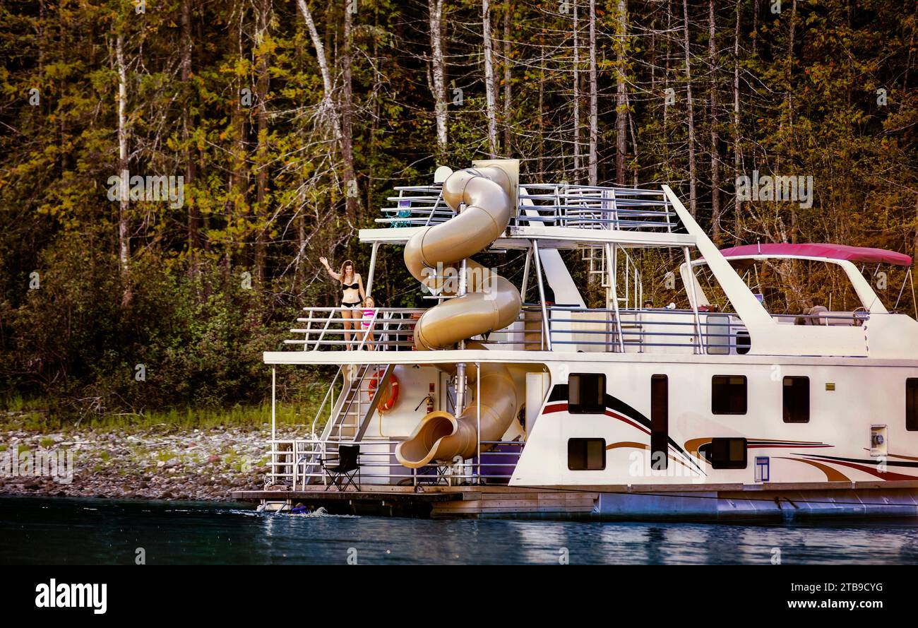 Una famiglia che si gode una vacanza in casa galleggiante mentre parcheggia sulla riva del lago Shuswap con mamma e figlia che saltano dal ponte a poppa del boa... Foto Stock