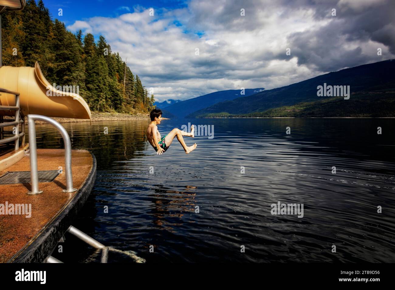 Ragazzo che salta dalla fine di uno scivolo acquatico su una casa galleggiante in un giorno autunnale sul lago Shuswap; British Columbia, Canada Foto Stock