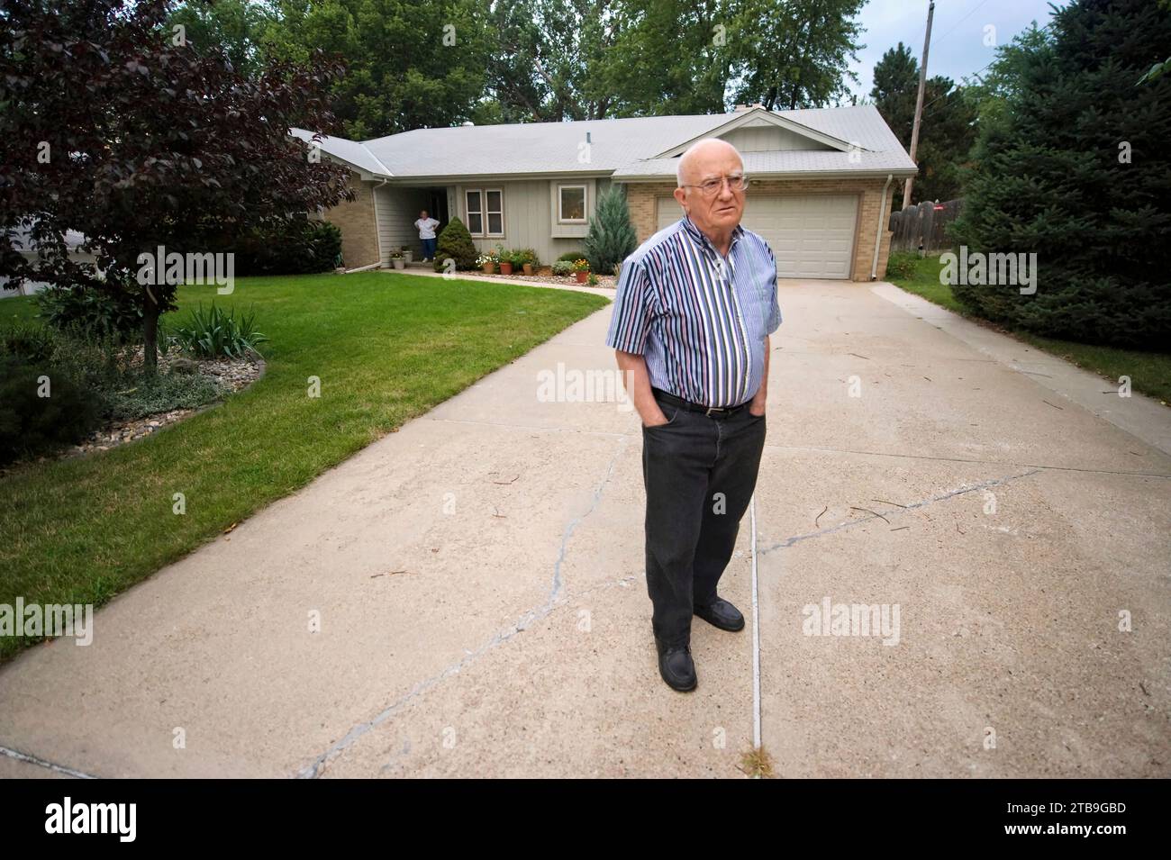 Un uomo anziano si trova nel vialetto di casa sua, con sua moglie in piedi davanti a doo correre in lontananza; Elkhorn, Nebraska, Stati Uniti d'America Foto Stock