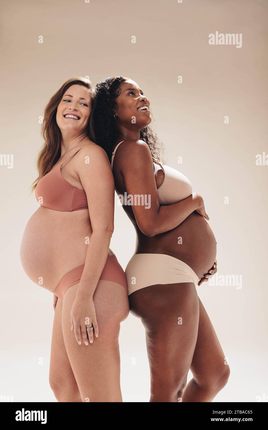 le donne incinte celebrano il loro corpo e abbracciano il loro viaggio di gravidanza in questa gioiosa immagine di studio. Due amici stanno insieme con i piccoli bump prou Foto Stock