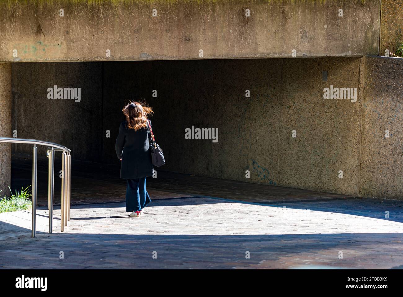 Una donna con la luce del sole che mette in risalto i capelli, cammina dalla luce del sole alle ombre sotto un tunnel in cemento a Sydney, Australia Foto Stock