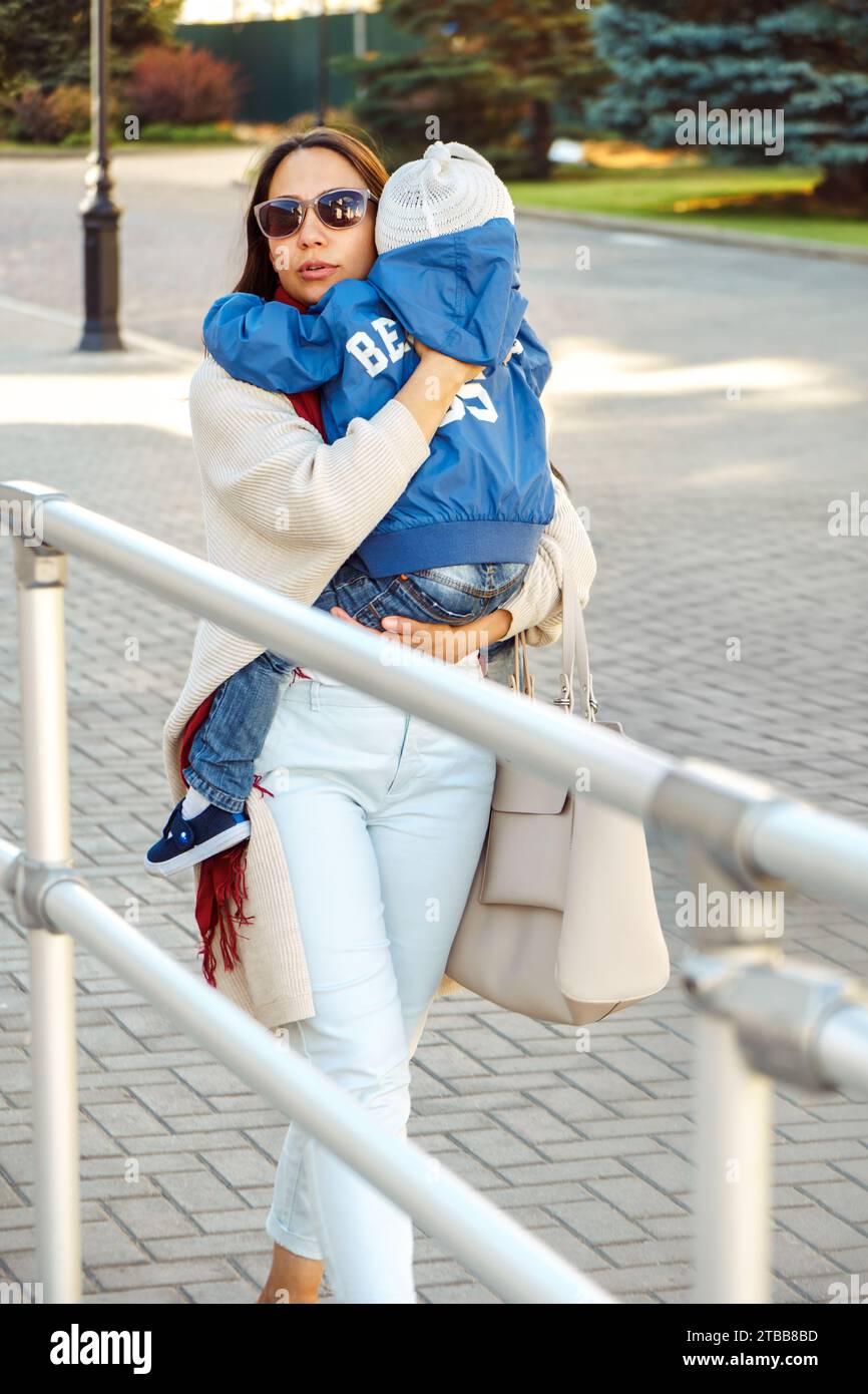 Una madre premurosa conforta il bambino che piange a spalla sulla strada cittadina. La donna porta il bambino arrabbiato a camminare lungo la piazza della città. Lady calma il bambino Foto Stock