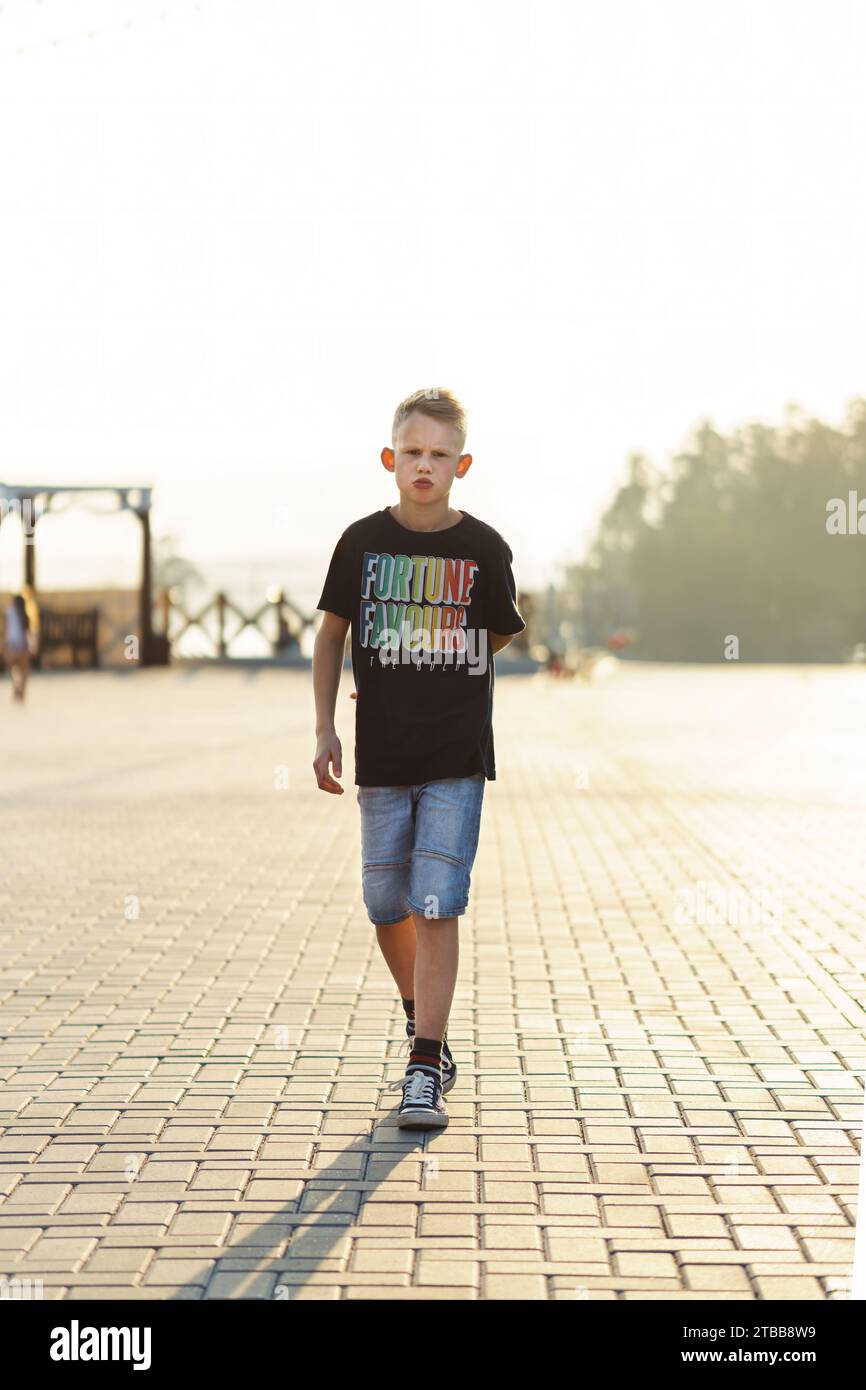 Un ragazzo in preda all'abbigliamento informale cammina lungo la piazza pavimentata della città. Il bambino trascorre il tempo a passeggiare sul terrapieno urbano alla luce del tramonto. Figo bambino Foto Stock