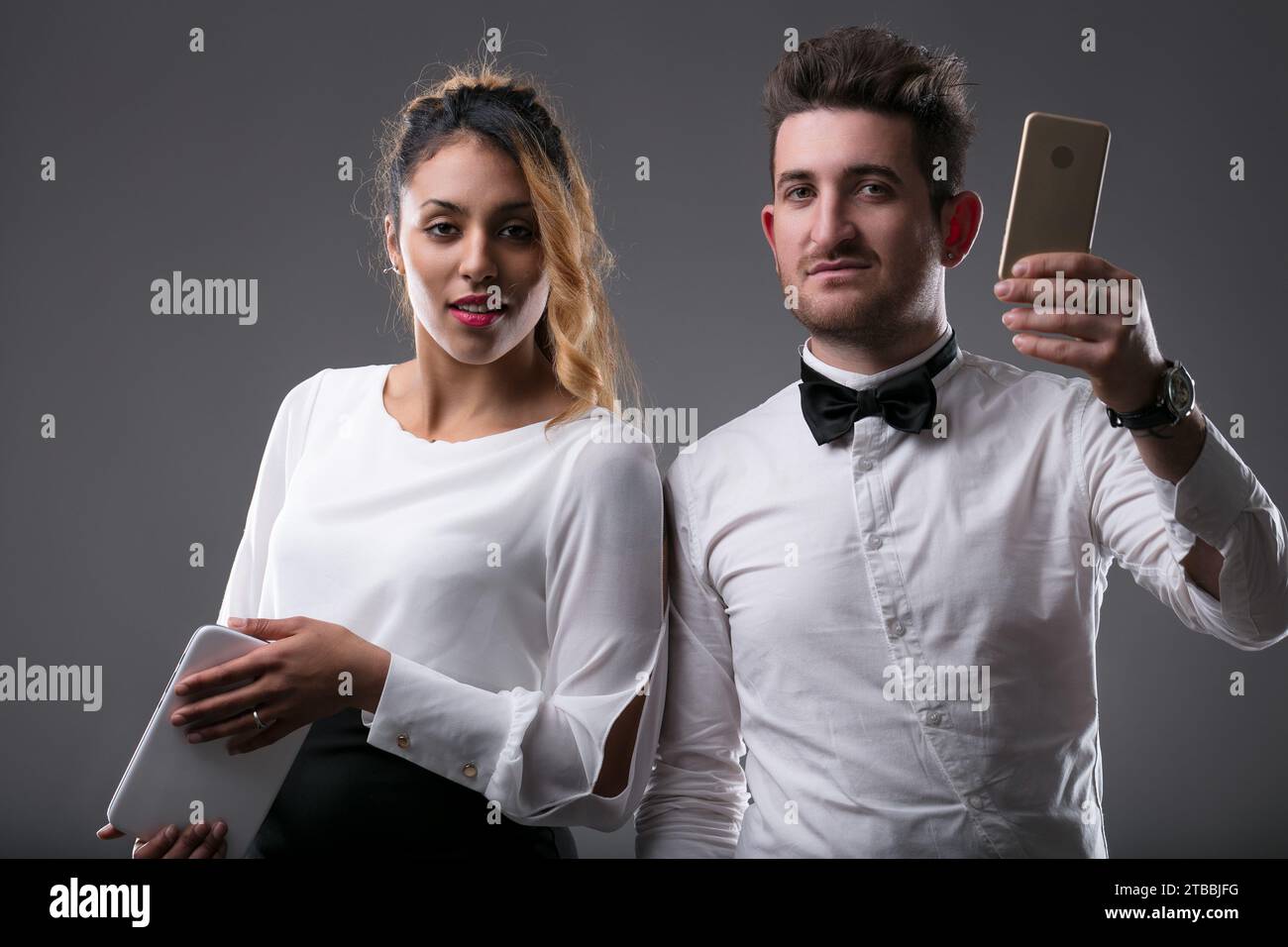 Donna elegantemente vestita si concentra su un tablet, l'uomo controlla il suo telefono Foto Stock