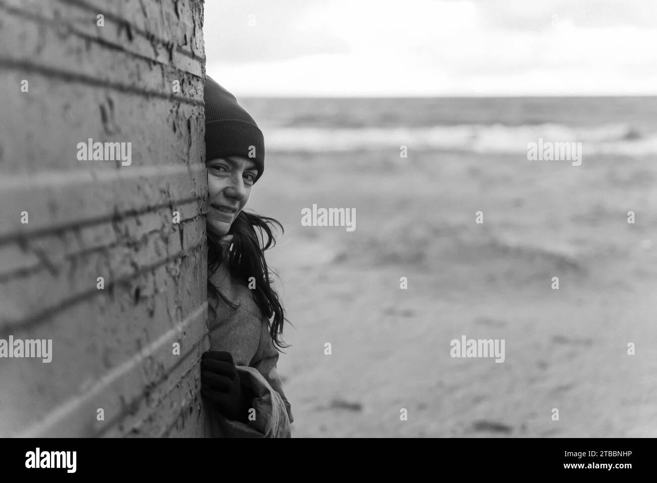 una donna con una giacca autunnale e un cappello rosso cammina lungo una spiaggia autunnale e posa accanto a un vecchio spogliatoio. foto in bianco e nero Foto Stock