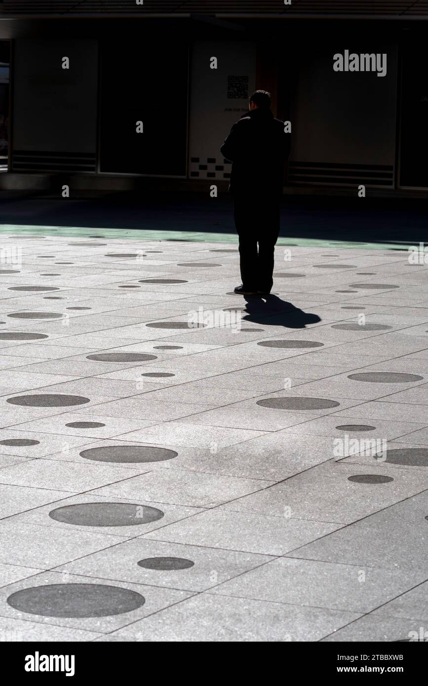 Un'immagine ad alto contrasto di un uomo in abiti neri, che cammina dalla luce all'ombra attraverso un'area pavimentata in cemento al Sydney Plaza in Australia Foto Stock