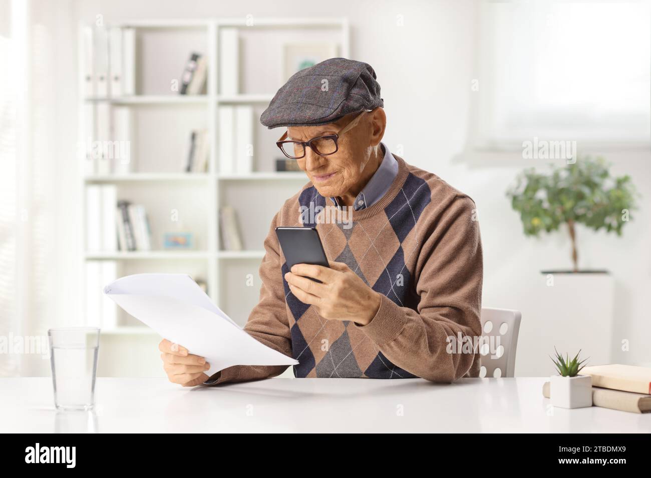 Un anziano a casa che ha uno smartphone e guarda un documento cartaceo Foto Stock