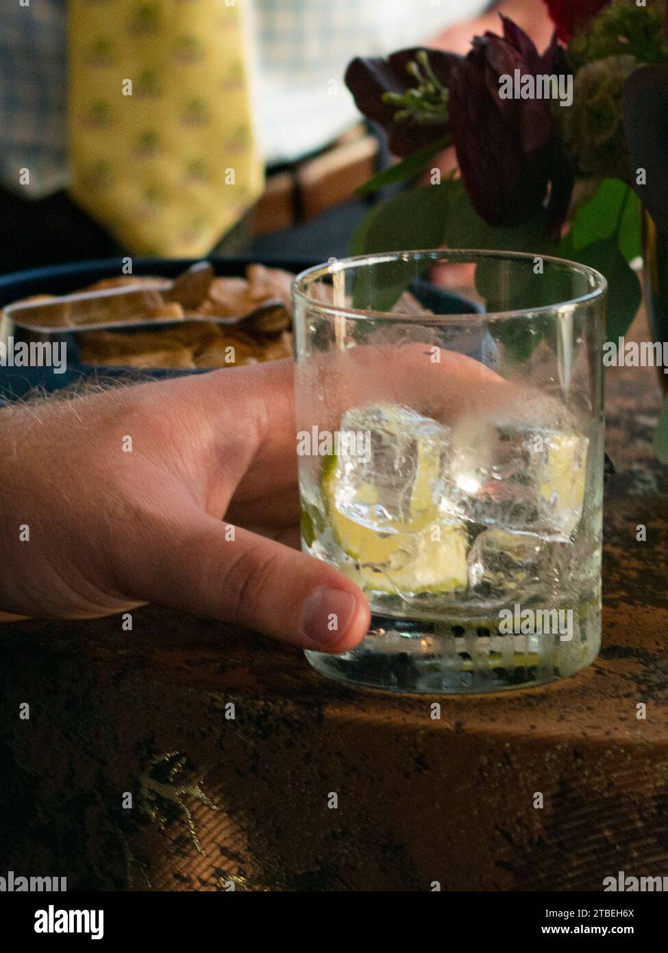 La mano dell'uomo regge un bicchiere da cocktail con cubetti di ghiaccio e cuneo di lime sul bordo del tavolo Foto Stock
