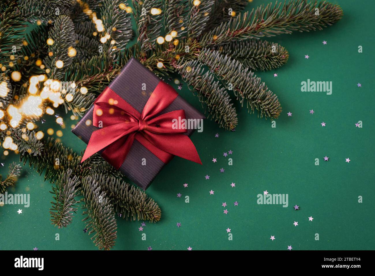 Confezione regalo con fiocco rosso e rami di abete su sfondo verde scuro con coriandoli. Vista dall'alto, base piatta, spazio di copia. Foto Stock