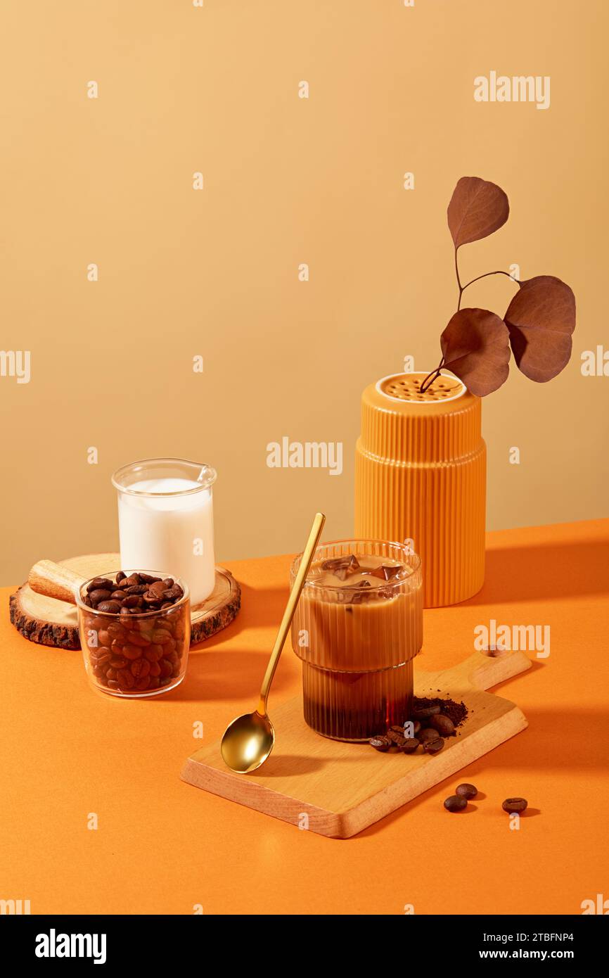 Tazza di caffè con latte e chicchi di caffè su tagliere di legno, circondata da oggetti di scena con sfondo marrone-arancio. I chicchi di caffè hanno molti benefici per la salute. Foto Stock