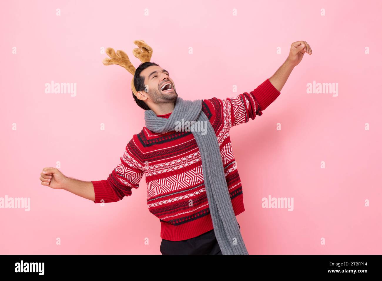 Ritratto in studio natalizio di un allegro uomo caucasico che indossa maglione e un elegante cerchietto di renna che ride e balla in uno sfondo isolato di colore rosa Foto Stock