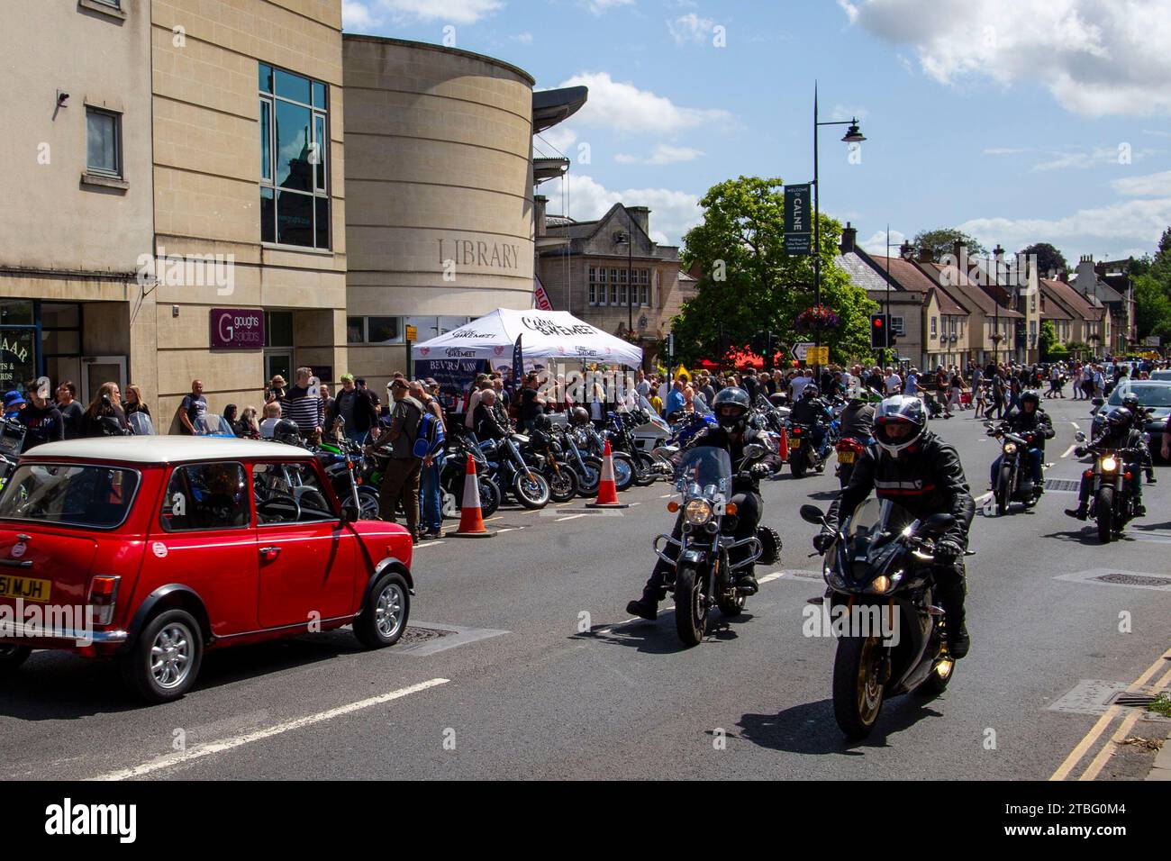 I motociclisti percorrono una strada trafficata costeggiata da spettatori e biciclette parcheggiate durante un evento pubblico vicino a un moderno edificio della biblioteca Calne bike Meet Foto Stock