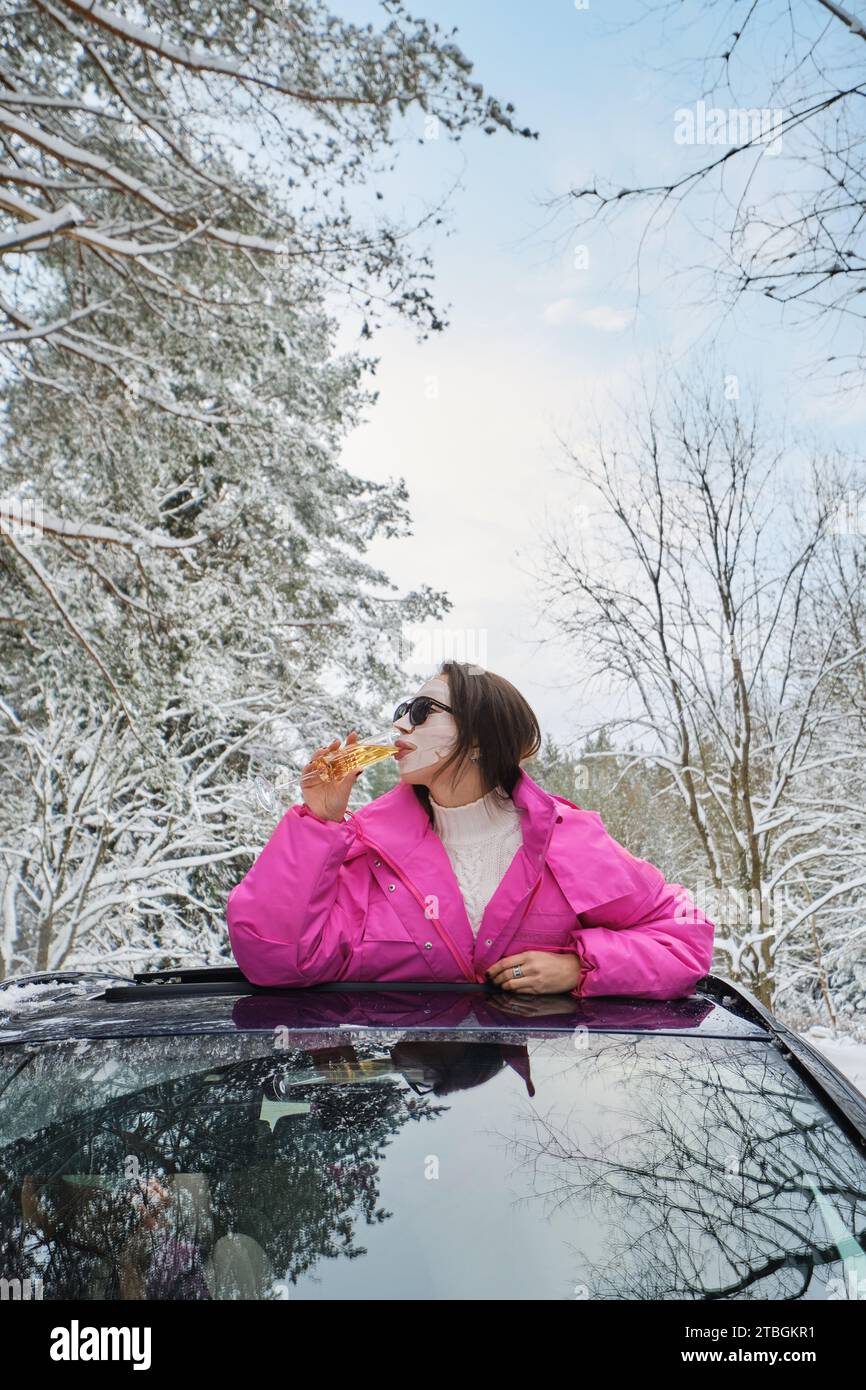 Una donna divertente che beve champagne che esce dal portello dell'auto nei boschi invernali Foto Stock