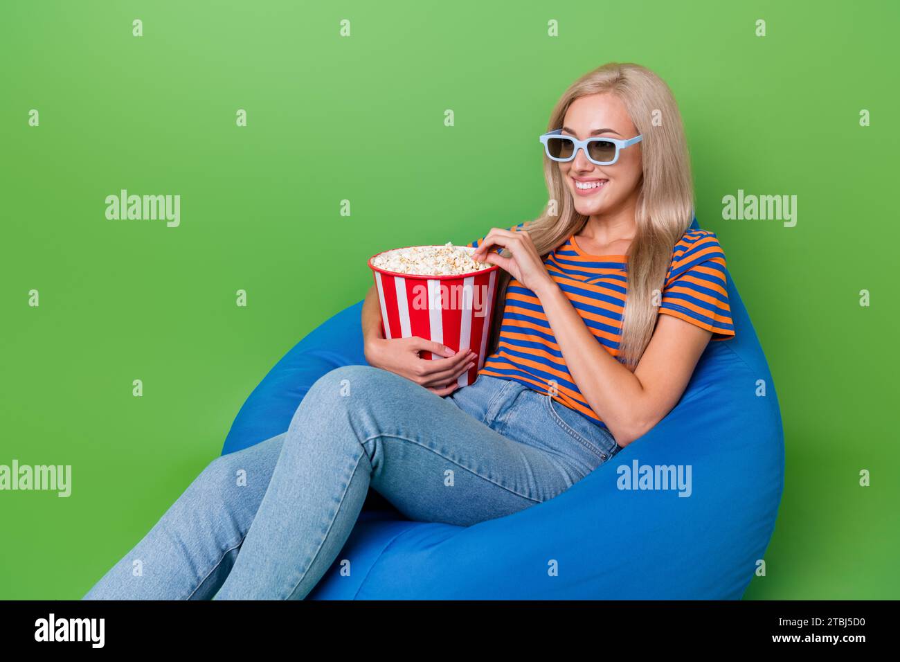 Foto di una ragazza felice che mangia il delizioso secchiello di popcorn salato preferito, che indossa un sacco di fagioli 3d isolato su sfondo verde Foto Stock