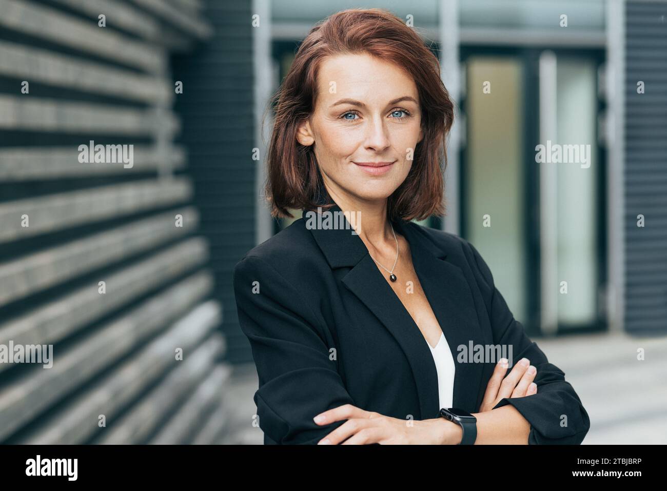 Donna sicura di mezza età con le braccia incrociate che guarda la fotocamera. Donna in abiti formali neri in posa all'aperto. Foto Stock