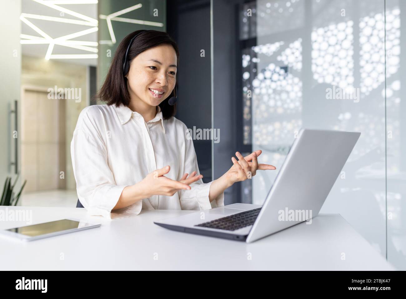 Donna d'affari sorridente di successo che lavora all'interno dell'ufficio sul posto di lavoro, che usa il laptop per videochiamare, sorridendo. Una donna asiatica utilizza una cuffia per consultare e comunicare con un cliente. Foto Stock