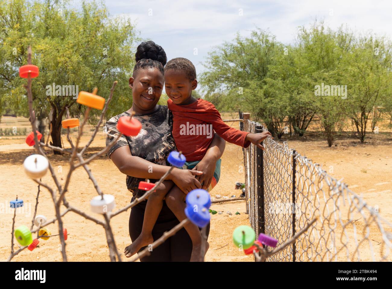 villaggio, donna africana che tiene il bambino davanti a un albero artistico con tappi colorati Foto Stock