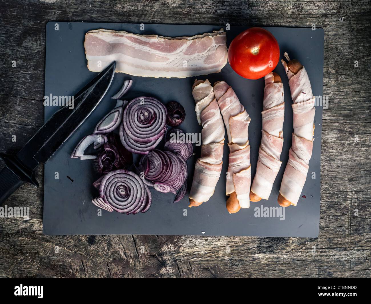 Piatto di salsicce ricoperte di pancetta, hot dog con cipolla di pomodoro e coltello da caccia, preparazione del cibo da picnic alla griglia all'aperto Foto Stock