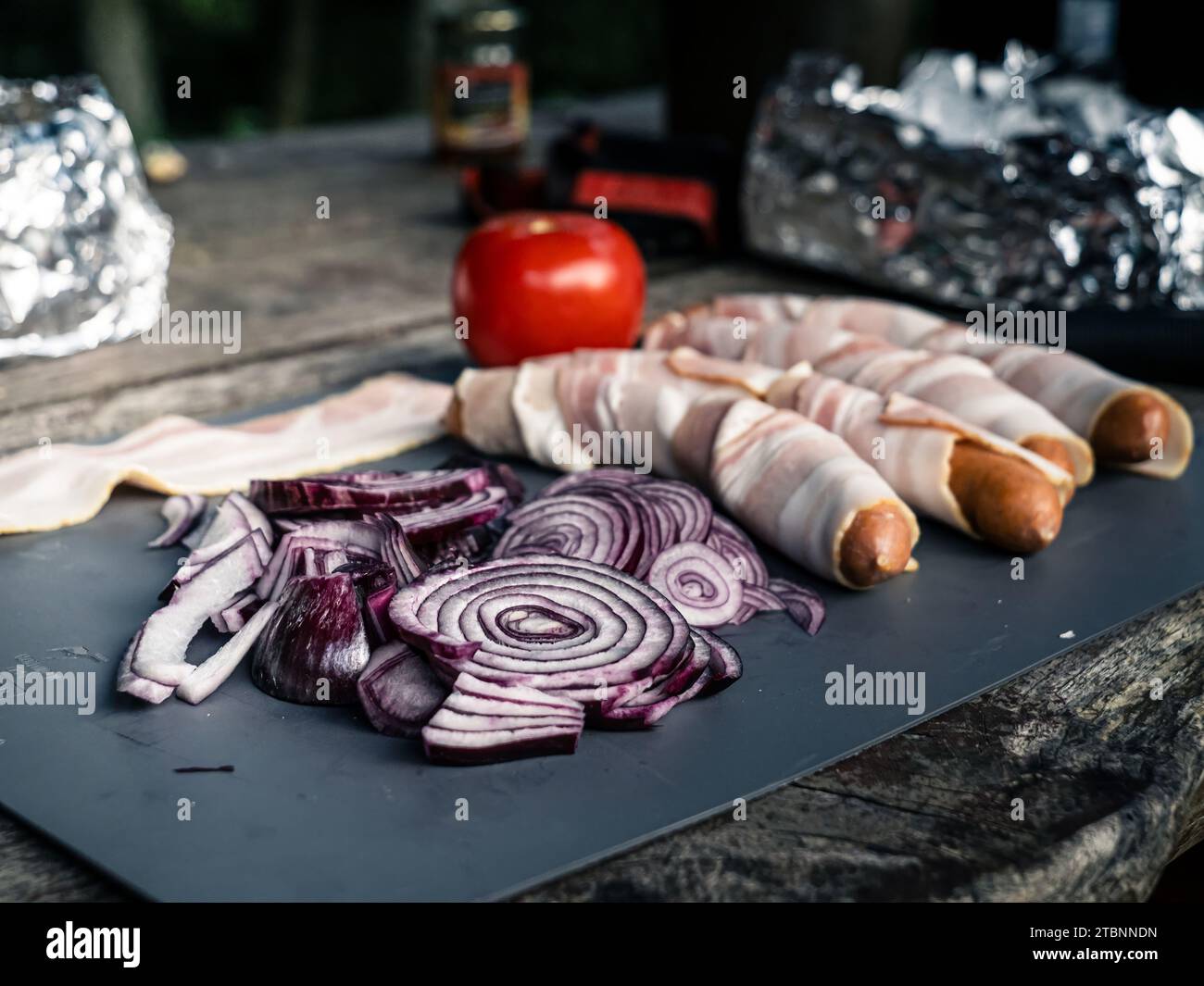 Foto di salsicce ricoperte di pancetta, hot dog con cipolla di pomodoro e un coltello da caccia, preparazione del cibo da picnic alla griglia all'aperto Foto Stock
