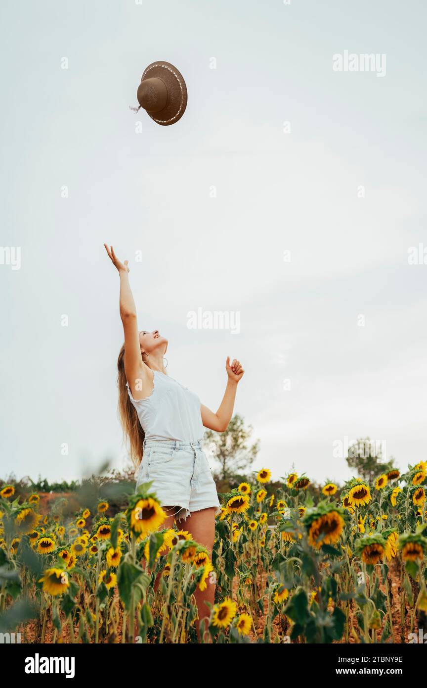 Giovane ragazza d'estate che lancia un cappello in aria circondata da fiori. Foto Stock