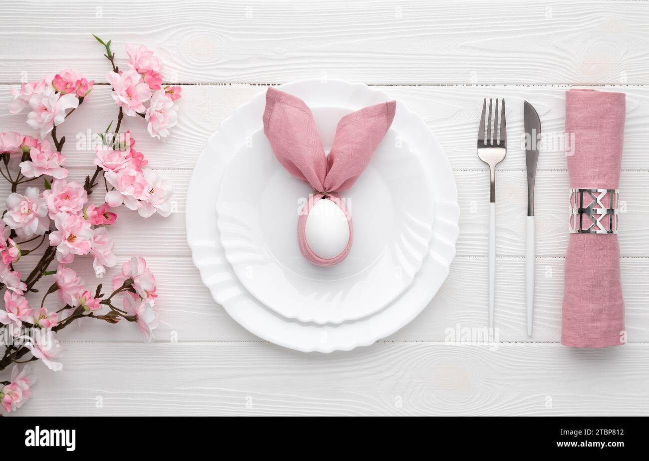 Tavolo per le festività pasquali con uovo su piatto e posate, fiori sakura e fiori rosa primaverili. Sfondo di legno bianco. Vista dall'alto, base piatta. Pranzo o. Foto Stock
