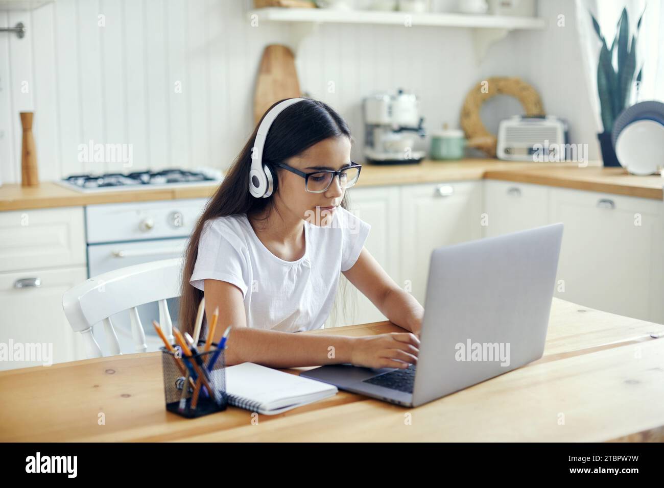 Una ragazza pre-adolescente di 12 anni con cuffie e occhiali si siede al tavolo e-learning, ascolta corsi online, lezioni audio, acquisire nuove conoscenze, competenze utilizzando Internet e. Foto Stock