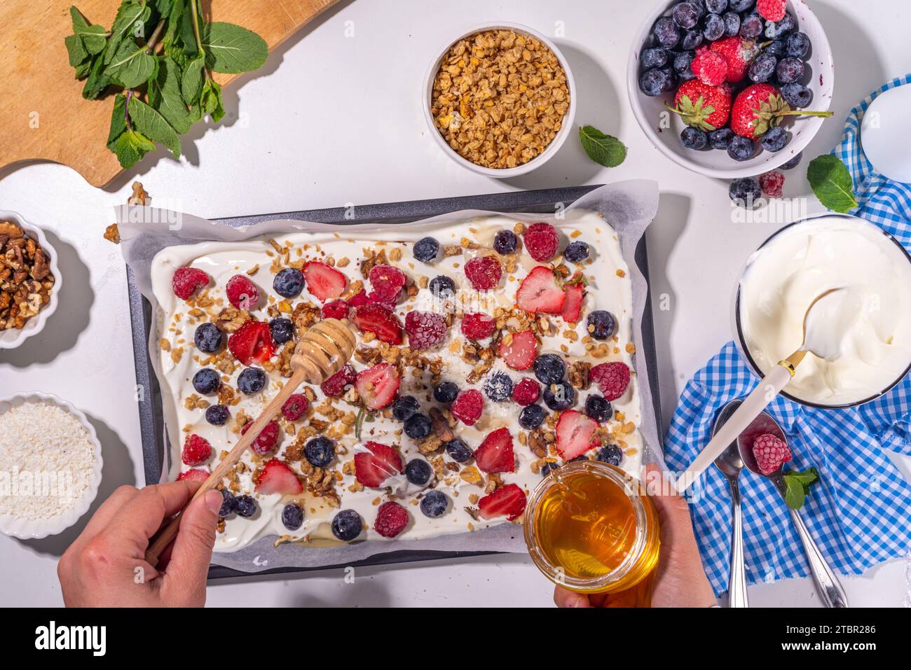 Preparazione della cottura con sfondo di corteccia di yogurt congelato con yogurt greco, bacche fresche, muesli, miele, salsa al cioccolato, con vassoio da forno deco. Vista dall'alto della donna Foto Stock