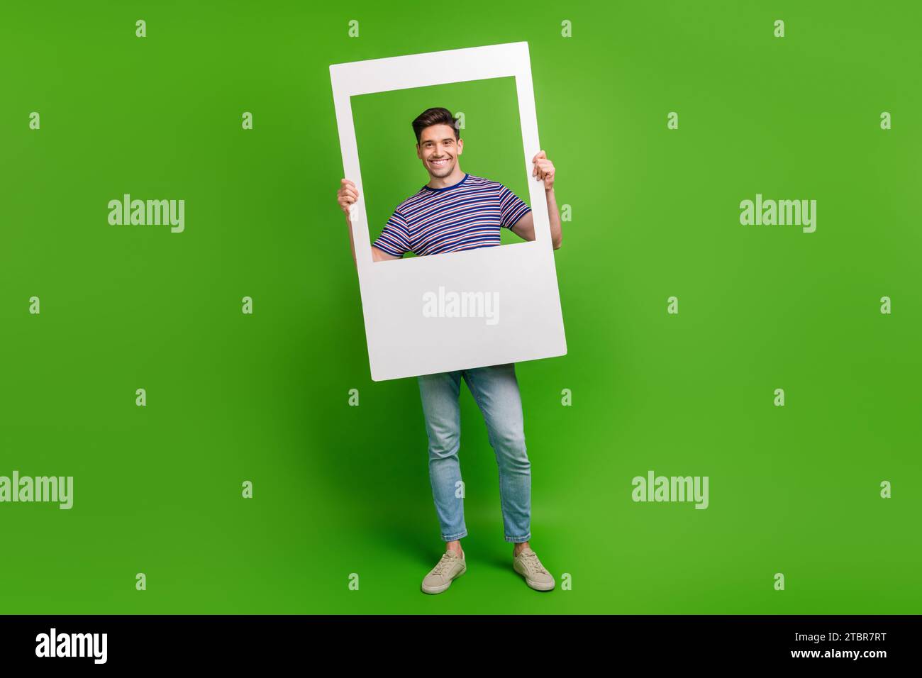 Foto a grandezza naturale di bellissimi pantaloni in denim vestiti da ragazzo di buon umore, camicia a righe che posa in una cornice fotografica isolata su sfondo di colore verde Foto Stock