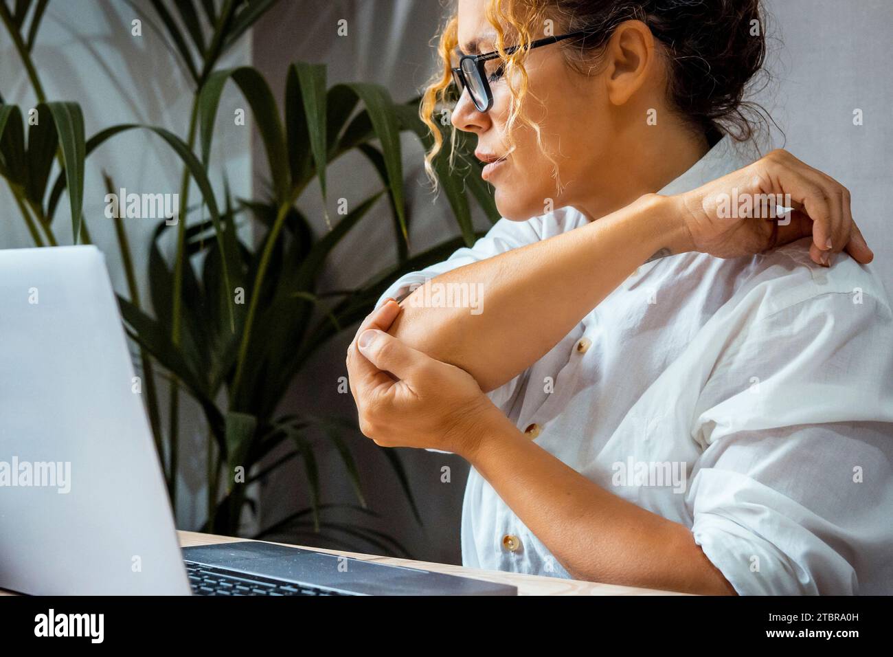Primo piano di una donna adulta che massaggia il gomito per un lavoro eccessivo scrivendo sul computer seduto alla scrivania in ufficio a casa. Concetto di espressione dolorosa donne stanche di lavorare su un notebook. Affari Foto Stock