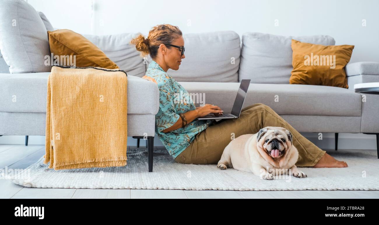 Vita domestica persone e animali insieme in amicizia e vita. Una donna che lavora con un computer portatile seduto sul tappeto a casa con il migliore amico cane che si rilassa vicino. Interno del soggiorno. Tempo libero. Foto Stock