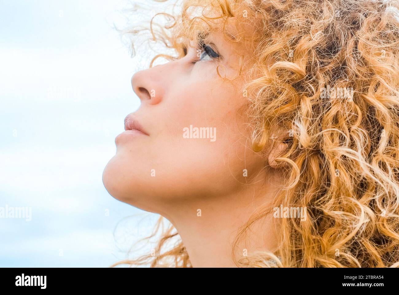 Primo piano di una bella donna di mezza età che guarda nell'aria. Capelli ricci naturali biondi. Bellezza femminile adulta. Una signora che pensa alle idee. Cielo di sfondo blu. Ritratti di persone ed espressioni. Foto Stock