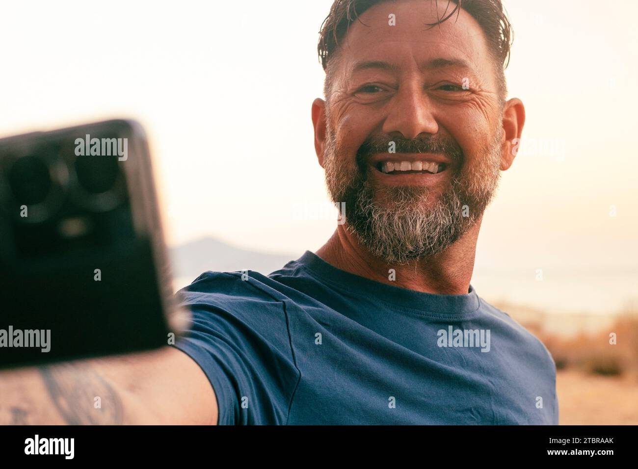 Uomo felice all'aperto che scatta foto selfie con lo smartphone mobile e sorride alla fotocamera. Condivisione online della vita sociale. Persone e foto di viaggio. Sfondo bianco del cielo paesaggio naturale, lato campagna Foto Stock