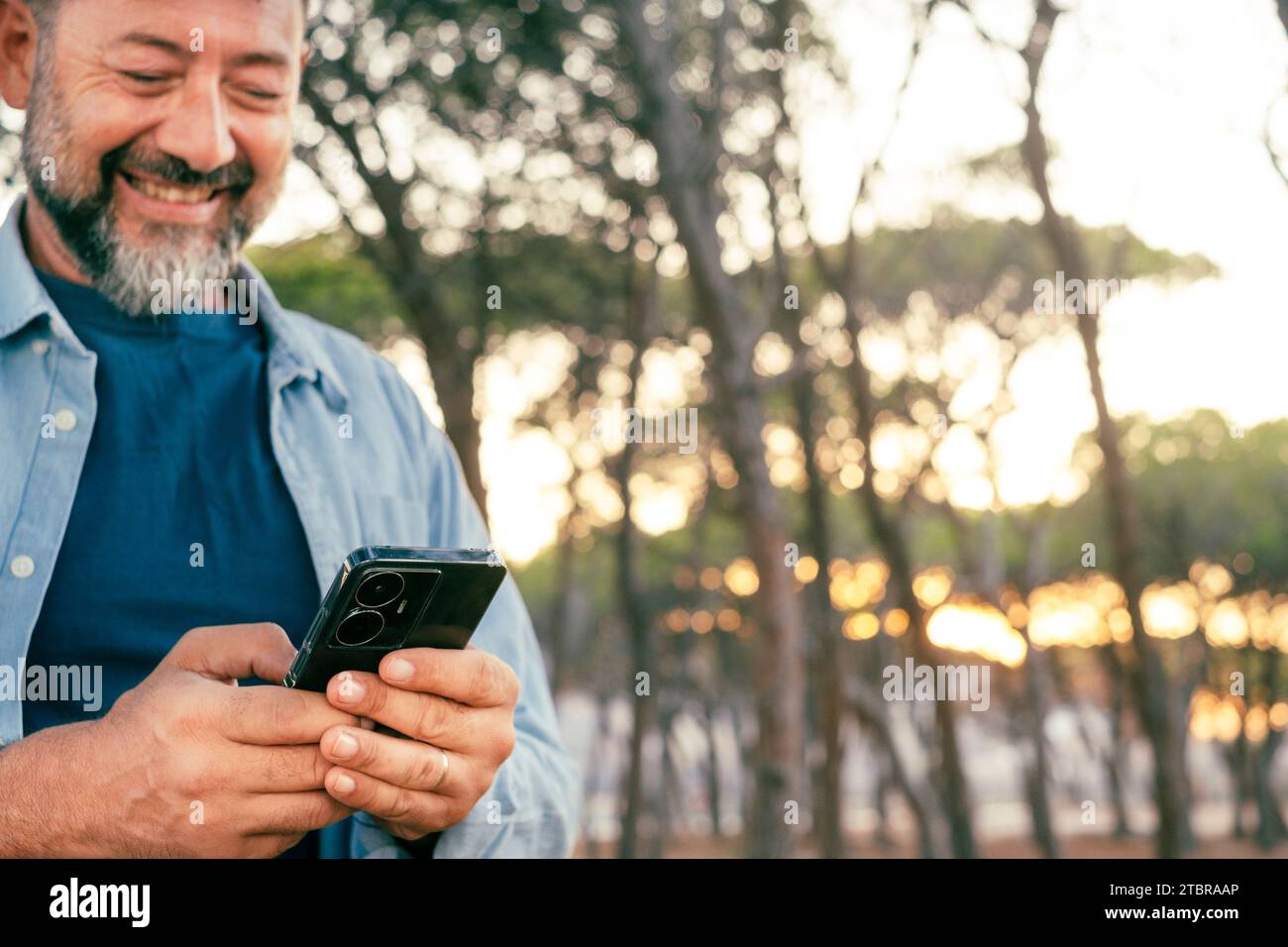 Un uomo moderno e maturo che usa uno smartphone all'aperto al parco sorridendo e divertendosi. Condivisione della vita sui social media caricamento di contenuti. Persone che scrivono sul cellulare in comunicazione esterna. Adulto Foto Stock