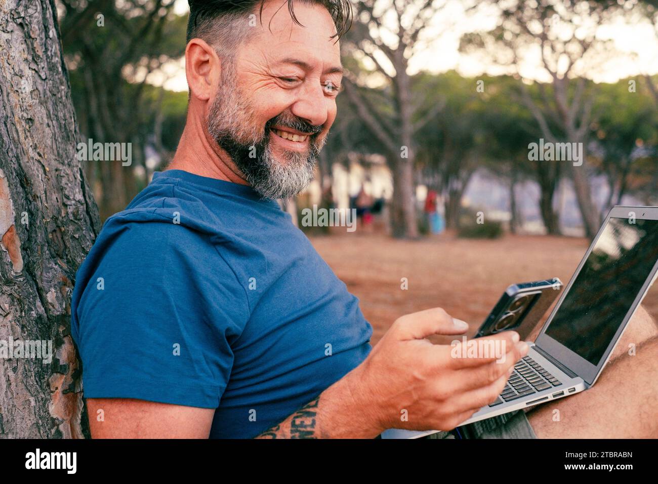 Un uomo adulto moderno che usa e lavora al computer portatile al parco. Concetto di smart working e stile di vita gratuito online per piccole e medie imprese nomadi digitali freelance Foto Stock