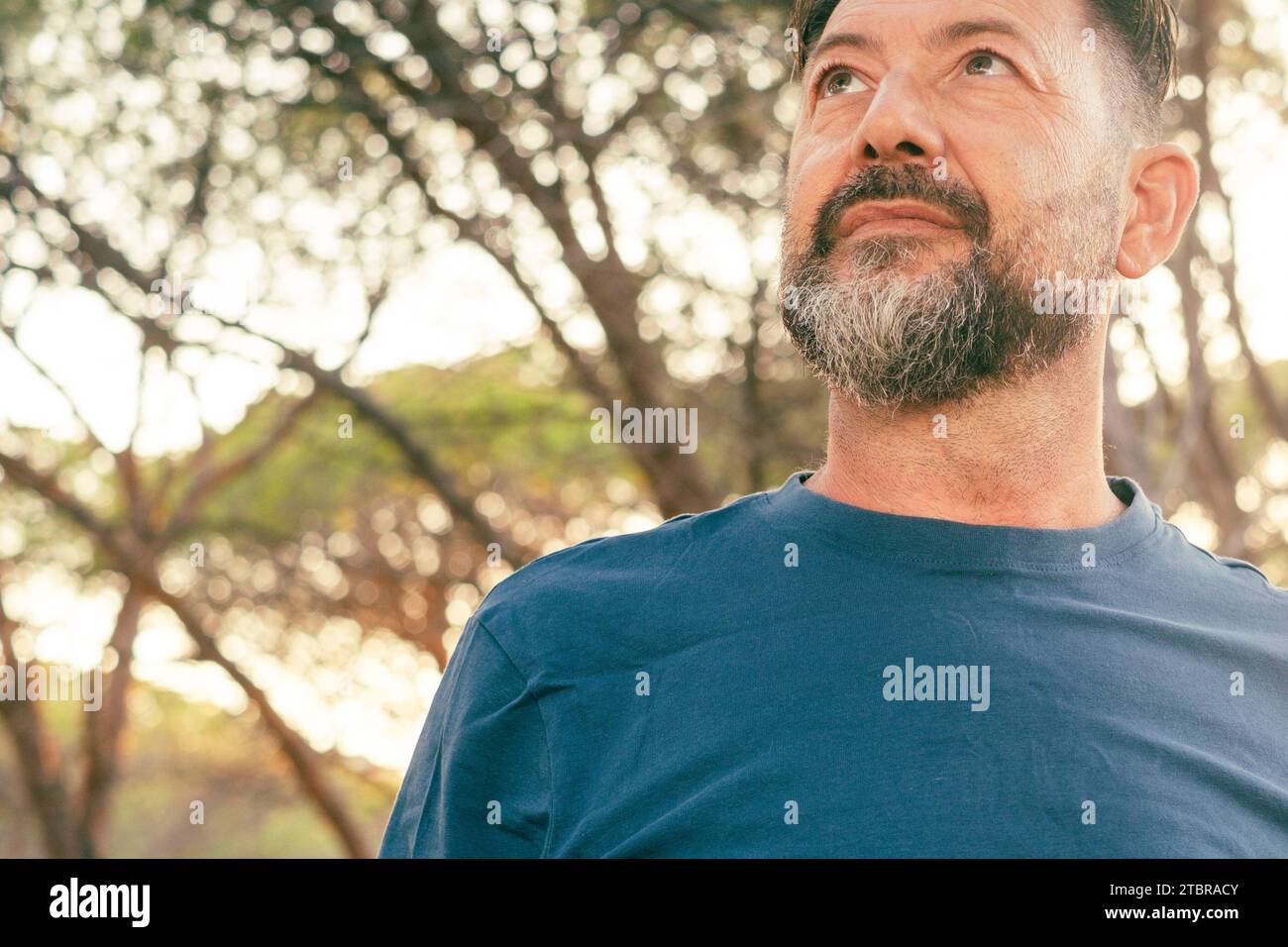 Ritratto ravvicinato di un uomo adulto sicuro di sé che guarda in alto. Uomo barbuto con maglietta blu e alberi sullo sfondo. Persone e attività ricreative all'aperto. Espressione serena sulla faccia. Copyspace Foto Stock