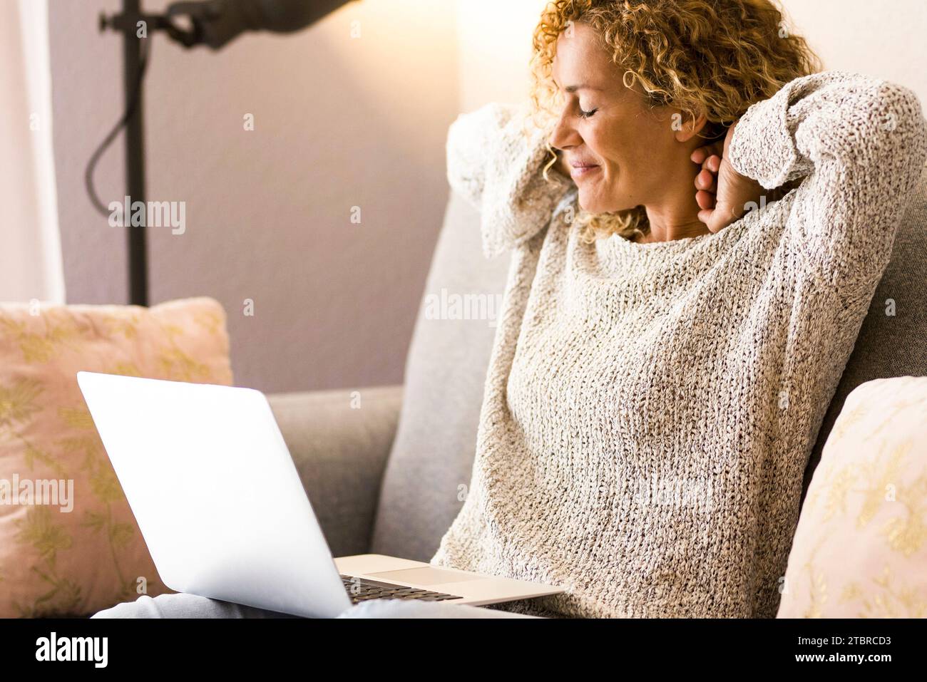 una donna finisce di usare il computer portatile a casa seduto sul divano allungando il collo e le spalle con un sorriso soddisfatto. Ritratto di una donna adulta moderna che usa un computer seduto sul divano. Interni. Foto Stock