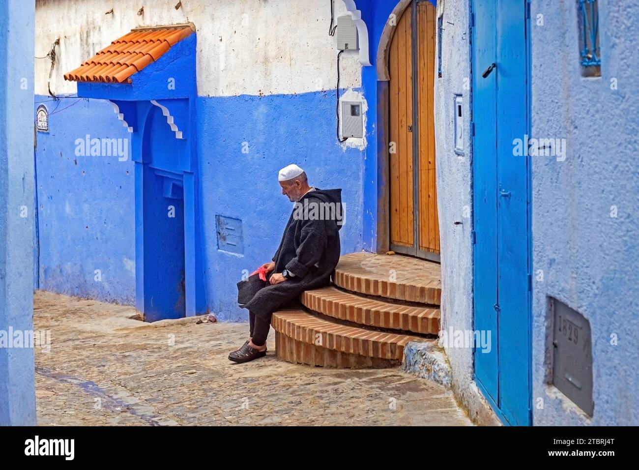 Uomo musulmano che indossa la tradizionale djellaba nel vicolo con case e porte blu nella medina / centro storico della città di Chefchaouen / Chaouen, Marocco Foto Stock