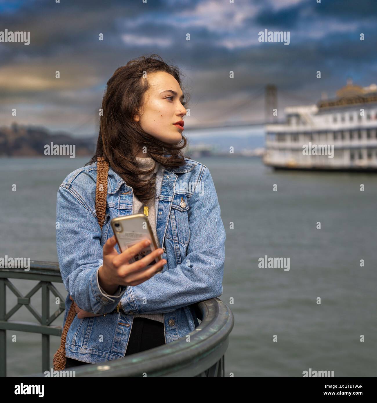 Unposed Teenage Female Waiting smartphone San Francisco Bay Oakland Bridge Cloudy Day Foto Stock