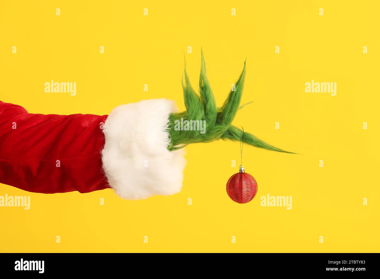 Mano pelosa verde di creatura in costume di Babbo Natale con pallina di Natale su sfondo giallo Foto Stock