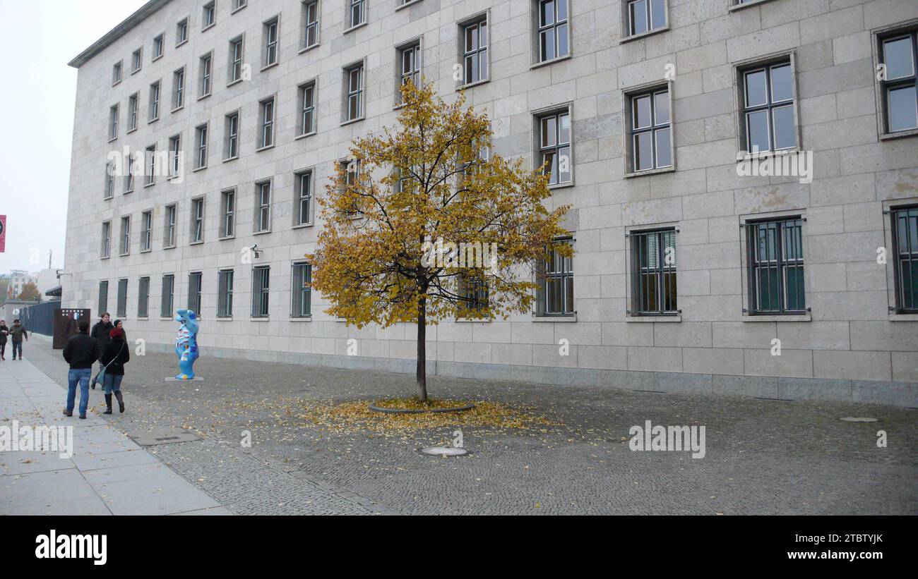 Un piccolo albero che lascia cadere foglie nella piazza di fronte all'edificio austero con telecamera di sicurezza, sculture di orsi di Berlino e diversi pedoni nelle vicinanze. Foto Stock