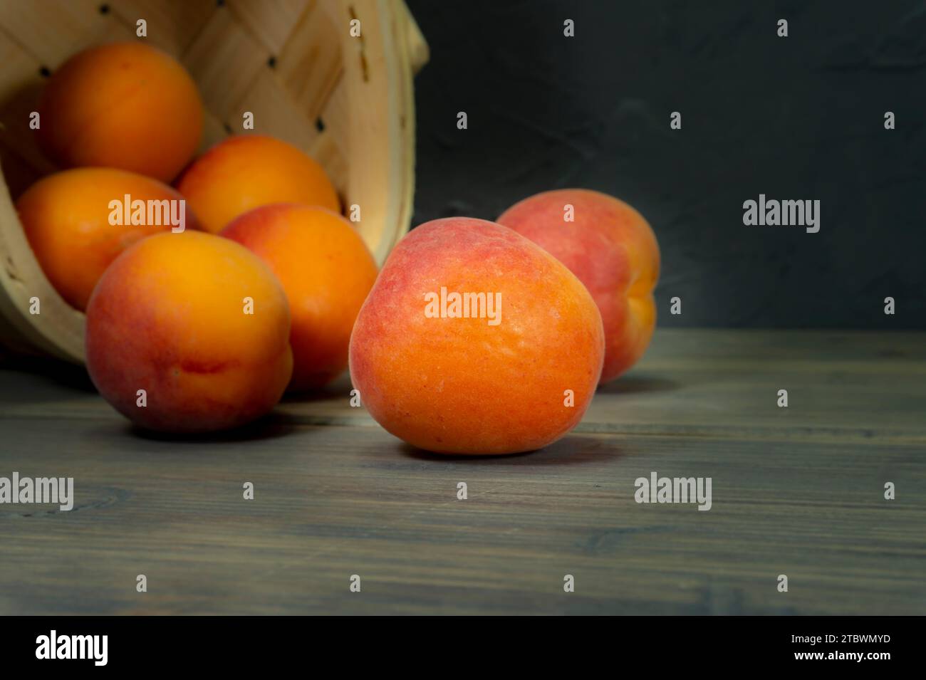 Pesche fresche che fuoriescono dal cestino di vimini ribaltato sul piano portapaziente in legno grigio. Muro nero sullo sfondo Foto Stock