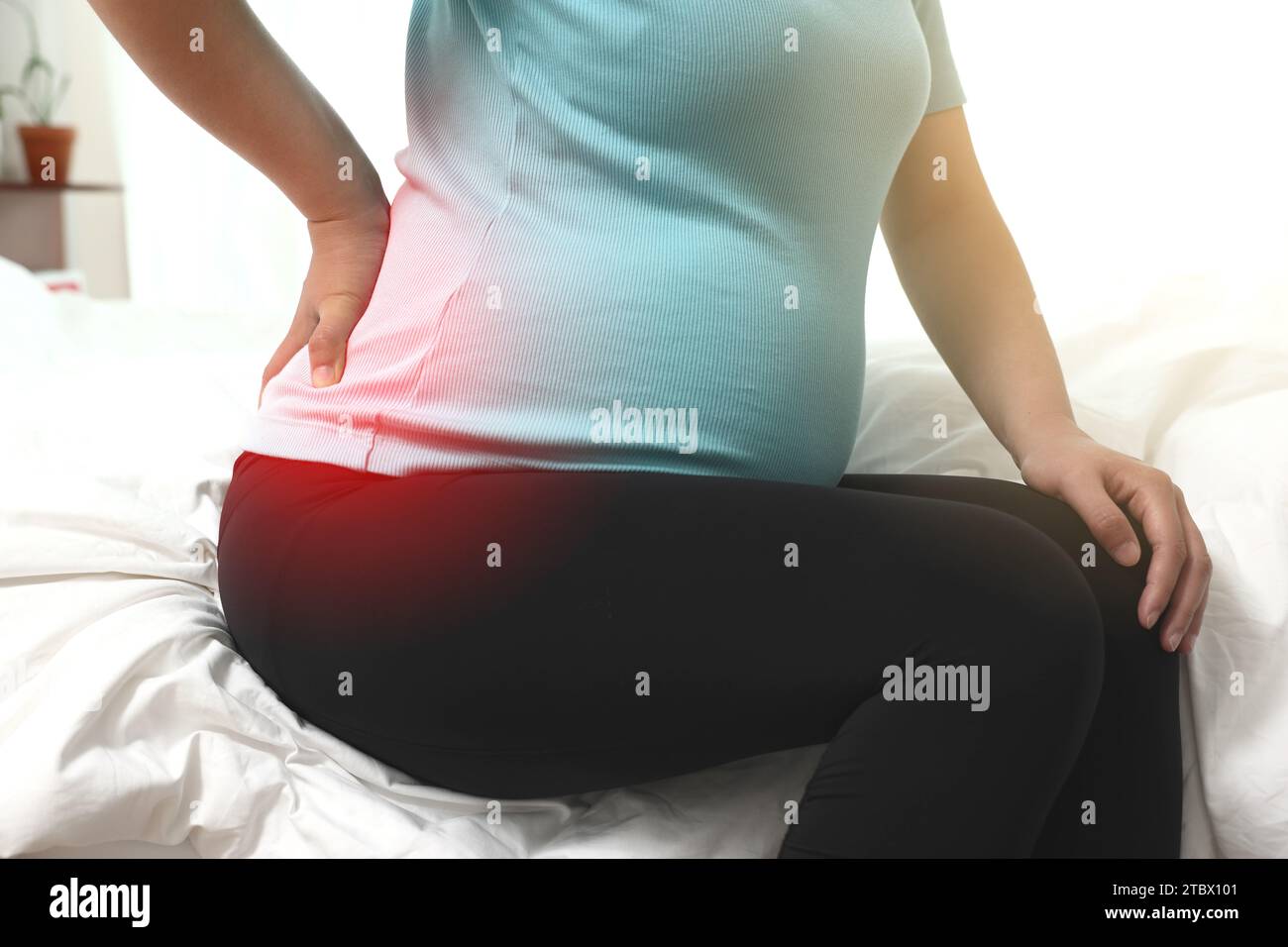 Una donna incinta è seduta su un letto toccando la schiena dolorante (evidenziata in rosso) Foto Stock