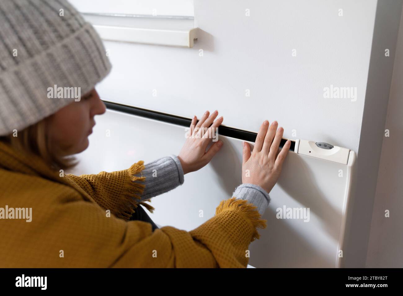 Una donna in abiti caldi tiene la mano sul riscaldatore. Bassa temperatura di riscaldamento in casa. Concetto di crisi energetica.. Foto Stock