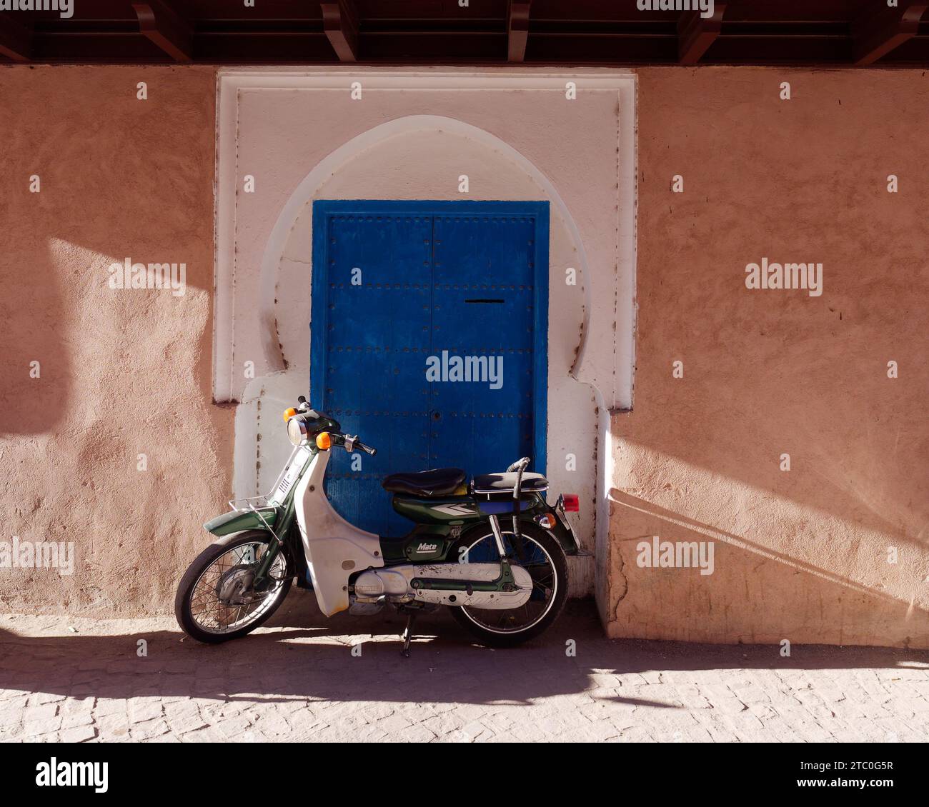 Motocicletta fuori da una porta blu incorniciata di bianco con una forte ombra nella Medina nella città di Marrakech, in Marocco, 9 dicembre 2023 Foto Stock