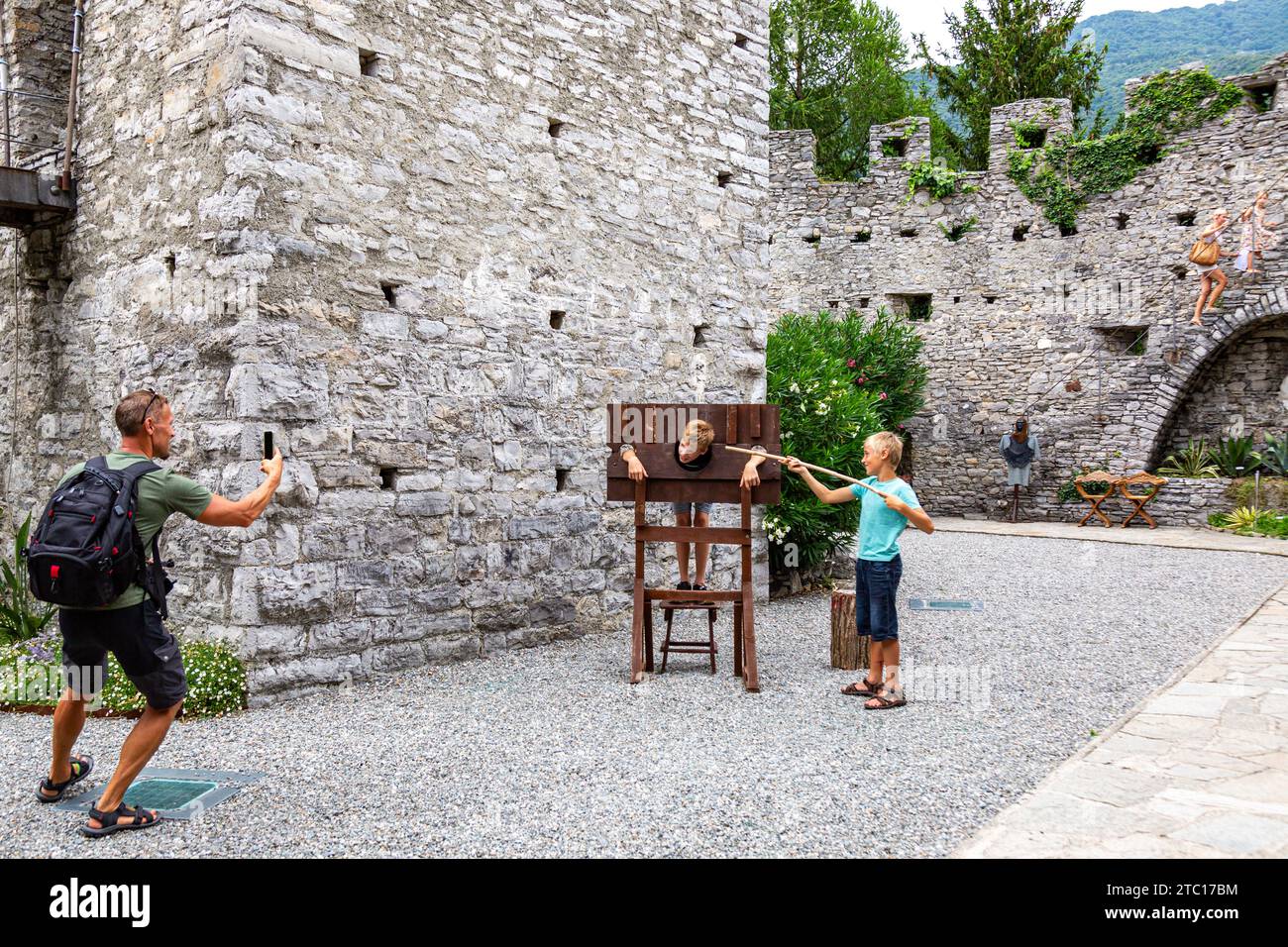 Un uomo fotografa i suoi figli come uno finge di essere prigioniero in una replica della gogna tra le rovine del medievale castello di Vezio a Perledo, Lombardia, Italia. Foto Stock