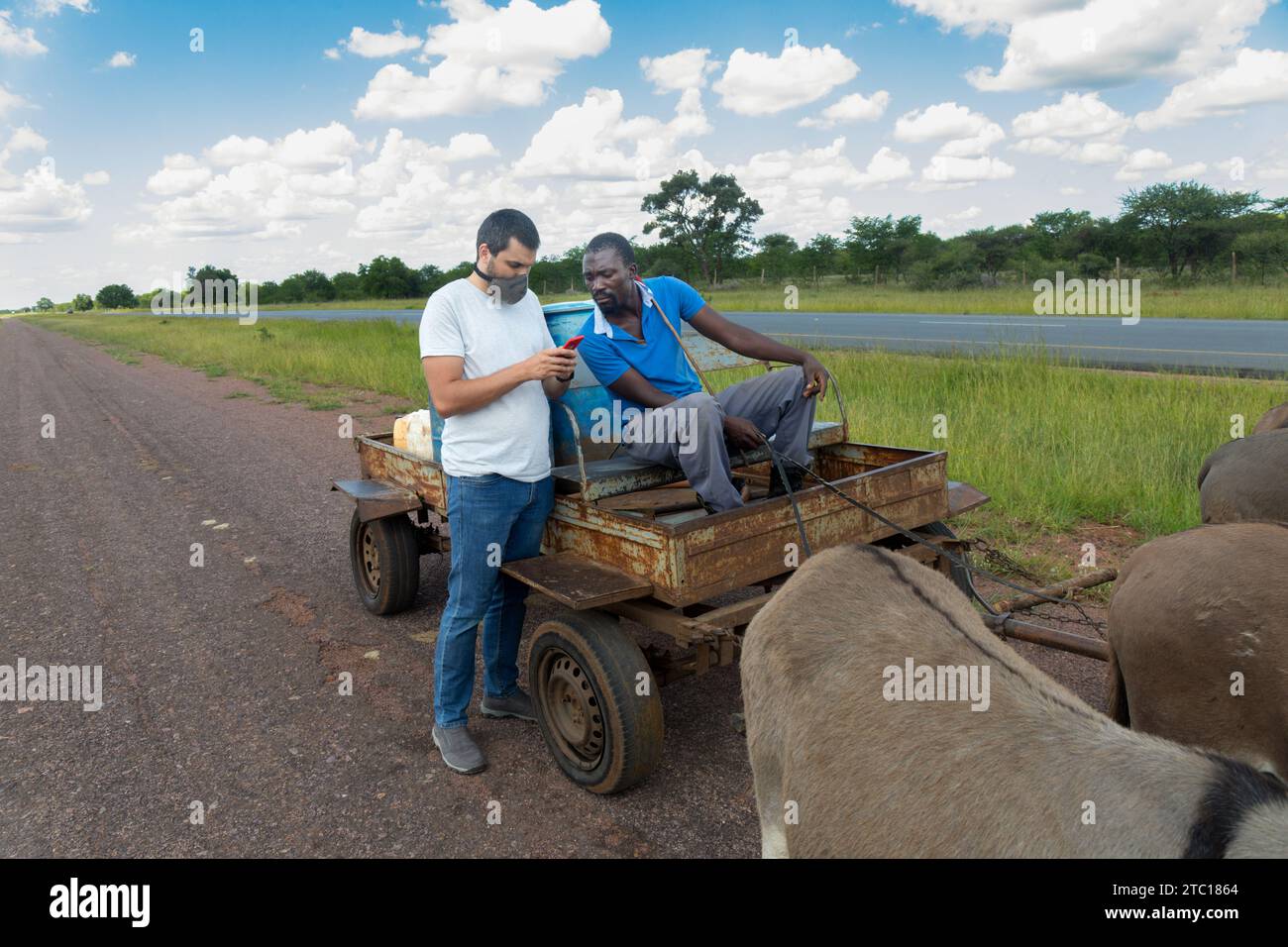 un uomo africano con un carrello d'asino che vende acqua nel villaggio, un uomo caucasico con il telefono che fa un accordo Foto Stock